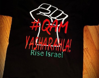 May include: A black short-sleeve t-shirt featuring a white graphic of a raised fist. The text #QAM YASHARAHLA! is printed in red, and the words Rise Israel are printed in green below the fist graphic on the front of the garment.