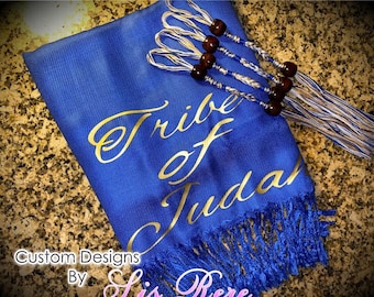May include: A royal blue prayer shawl featuring the gold text Tribe of Judah printed diagonally across the fabric. The shawl includes a fringe at the bottom and four decorative braided cords at the top, each adorned with dark brown wooden beads.