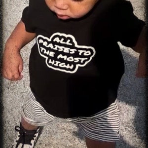 May include: A young child wearing a black t-shirt with the text "ALL PRAISES TO THE MOST HIGH" in white, paired with striped shorts and black and white sneakers. The image is framed with a dark border.