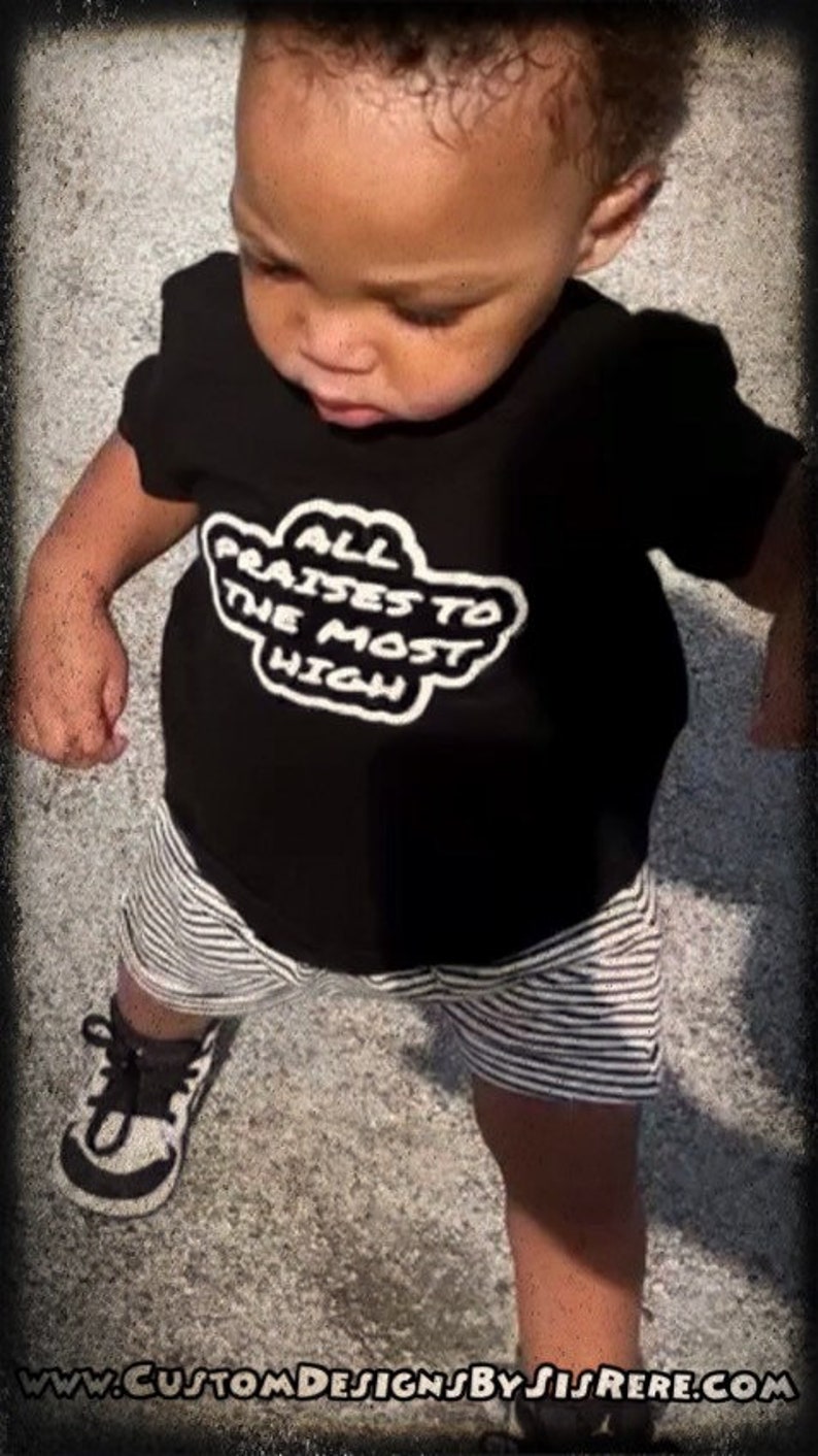 May include: A young child wearing a black t-shirt with the text "ALL PRAISES TO THE MOST HIGH" in white, paired with striped shorts and black and white sneakers. The image is framed with a dark border.