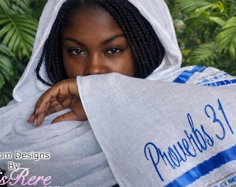May include: A white hooded scarf featuring blue ribbon trim and the embroidered text Proverbs 31 in blue script. The fabric has a lightweight texture, and the design includes a decorative border with blue and white patterns along the edge.