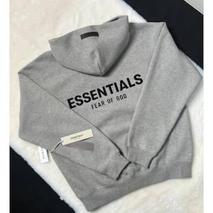 Puede incluir: Una sudadera con capucha de color gris jaspeado que presenta el texto negro ESSENTIALS FEAR OF GOD impreso en la espalda. La sudadera incluye un parche rectangular oscuro en la capucha y etiquetas de papel con la marca y el logo de PacSun.