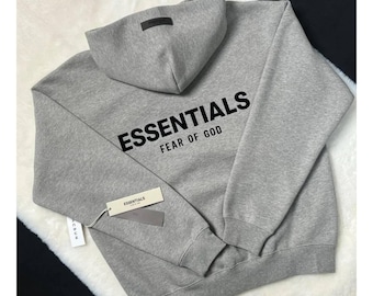 May include: A heather grey hooded sweatshirt featuring the black text ESSENTIALS FEAR OF GOD printed on the back. The hoodie includes a dark rectangular patch on the hood and attached paper tags with the brand name and PacSun branding.
