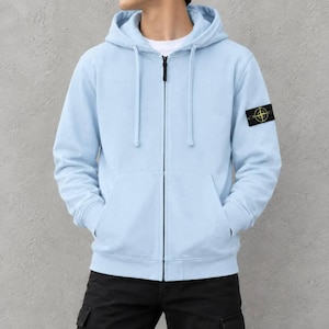 Op de afbeelding: Een lichtblauwe hoodie met ritssluiting, voorzien van een capuchon met trekkoord, lange mouwen en een zwarte patch op de linkermouw met een geel kompaslogo. Het kledingstuk heeft een volledige ritssluiting aan de voorzijde en twee zakken.