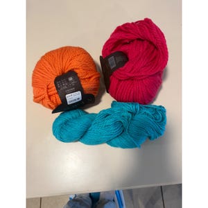 Könnte beinhalten: Drei Garnknäuel in leuchtendem Orange, kräftigem Pink und Türkisblau. Die orangefarbenen und pinken Knäuel haben schwarze Papieretiketten mit weißem Text, einschließlich des Markennamens Rowan und Pflegehinweisen, während das türkise Garn lose ist.