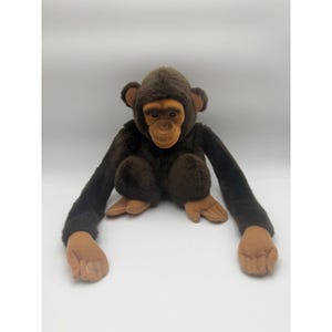 May include: A plush chimpanzee toy with dark brown fur and a light tan face, hands, and feet. The stuffed animal features long arms, rounded ears, and a soft, textured body, designed in a seated position with palms facing upward and forward.