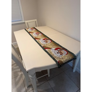 May include: A rectangular table runner featuring a repeating pattern of Santa Claus faces with white beards and red hats on a gold background with star accents, bordered by a dark green trim with the text "I believe in Santa Claus".