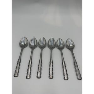May include: A set of six silver-toned metal spoons arranged in a row. Each spoon features a polished oval bowl and a slender handle decorated with a repeating embossed floral pattern along the edges, ending in a rounded tip with a subtle design.