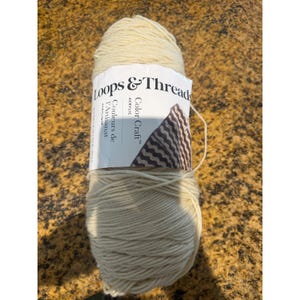 May include: A skein of off-white acrylic yarn with a white paper label featuring the text Loops & Threads Color Craft and Couleurs de l'Artisanat. The label displays a small graphic of a brown and white wavy pattern on the right side.