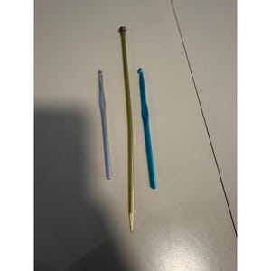 May include: Three metal crochet hooks of varying lengths and colors. The left hook is light purple, the center hook is a long gold-toned needle with a small cap at the top, and the right hook is bright blue. Each tool features a hook at one end.