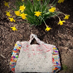 May include: A white crocheted tote bag with colorful plastic strips woven into the sides. The bag is sitting on a bed of brown mulch with yellow daffodils in the background.