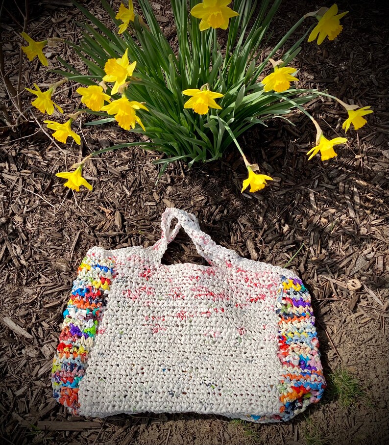 May include: A white crocheted tote bag with colorful plastic strips woven into the sides. The bag is sitting on a bed of brown mulch with yellow daffodils in the background.