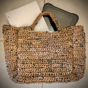 May include: A brown crocheted tote bag with a green and brown pattern. The bag is filled with a white sheet of paper and a black tablet computer.