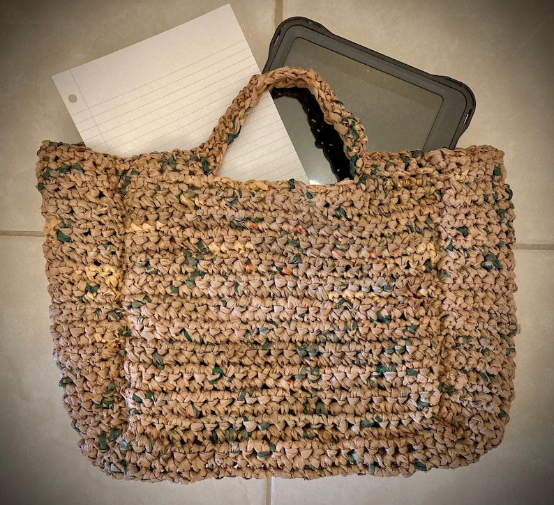 May include: A brown crocheted tote bag with a green and brown pattern. The bag is filled with a white sheet of paper and a black tablet computer.