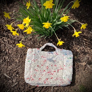 May include: A white crocheted tote bag with red and green accents. The bag is made from recycled plastic and is sitting on a bed of brown mulch. Yellow daffodils are in the background.