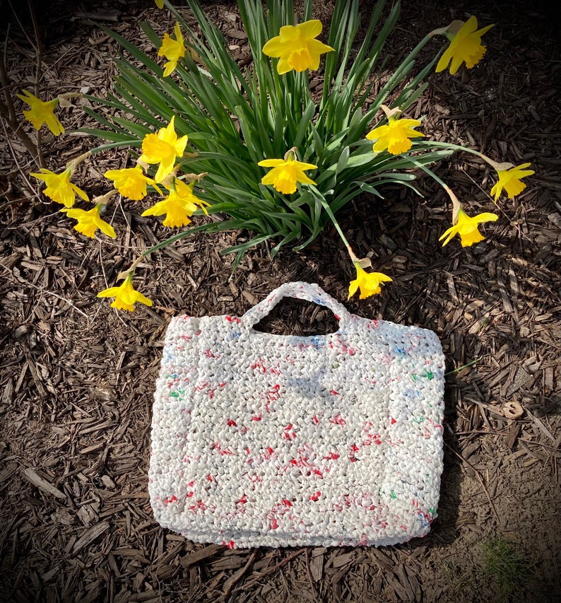 May include: A white crocheted tote bag with red and green accents. The bag is made from recycled plastic and is sitting on a bed of brown mulch. Yellow daffodils are in the background.