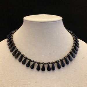 May include: A black beaded necklace with a teardrop pendant design. The necklace is displayed on a white mannequin bust against a black background. The teardrop pendants are evenly spaced and hang from a strand of small black beads.