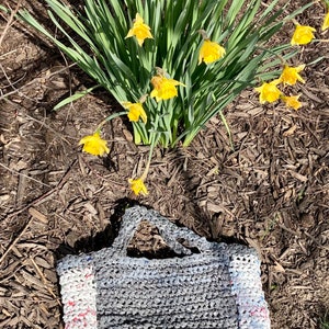 May include: A gray crocheted tote bag with white and red trim. The bag is made from recycled plastic and is sitting on a bed of brown mulch. Yellow daffodils are in the background.