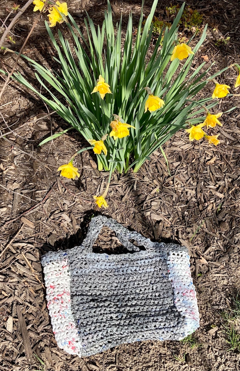 May include: A gray crocheted tote bag with white and red trim. The bag is made from recycled plastic and is sitting on a bed of brown mulch. Yellow daffodils are in the background.