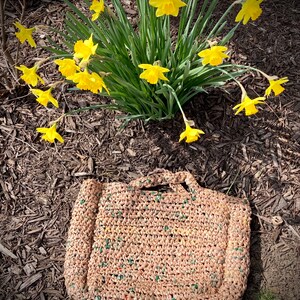 May include: A brown crocheted tote bag with green and white accents. The bag is sitting on a bed of brown mulch. There are yellow daffodils in the background.