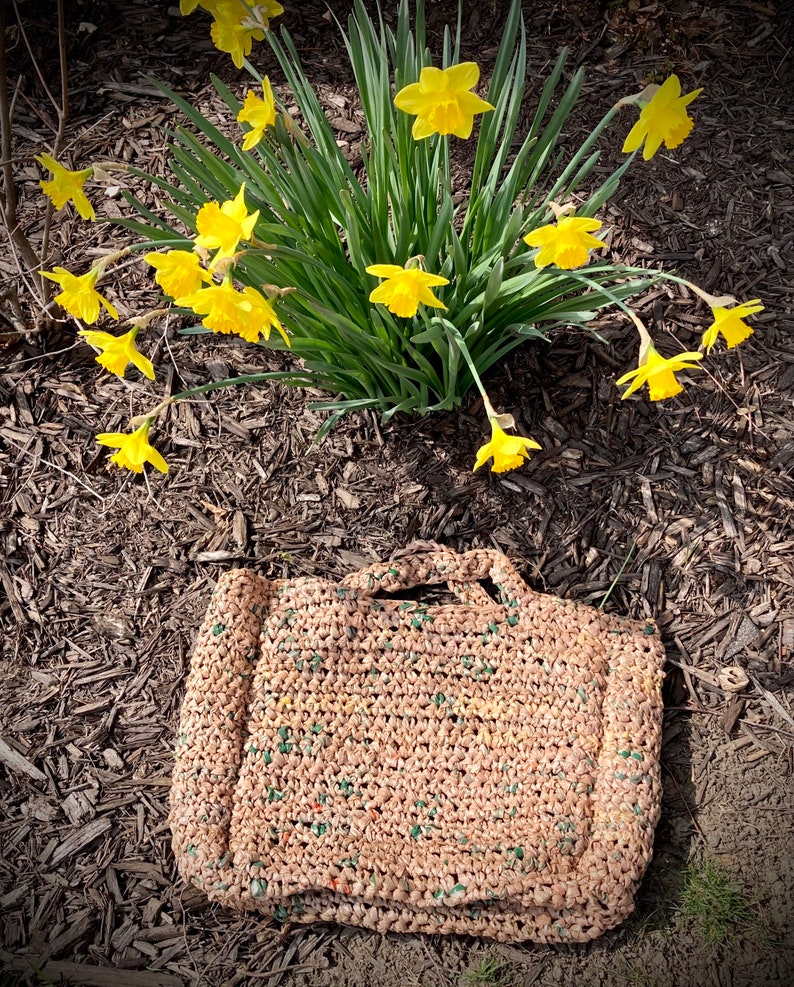 May include: A brown crocheted tote bag with green and white accents. The bag is sitting on a bed of brown mulch. There are yellow daffodils in the background.