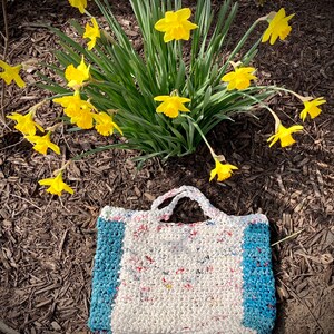 May include: A white crocheted tote bag with blue trim. The bag is made from recycled plastic and has a unique, textured look. The bag is sitting on a bed of brown mulch.