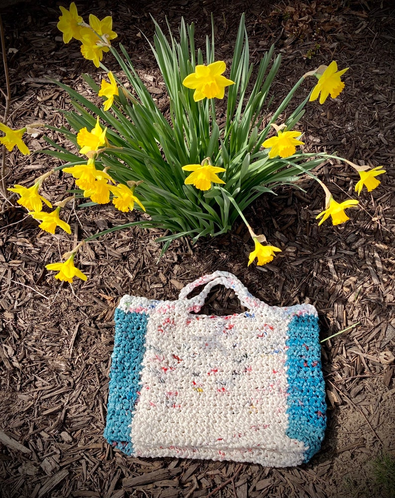 May include: A white crocheted tote bag with blue trim. The bag is made from recycled plastic and has a unique, textured look. The bag is sitting on a bed of brown mulch.