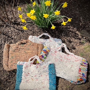 May include: Three crocheted bags made from recycled plastic. The bags are in different colors and sizes. The largest bag is white with colorful accents, the medium bag is brown, and the smallest bag is white with blue accents.
