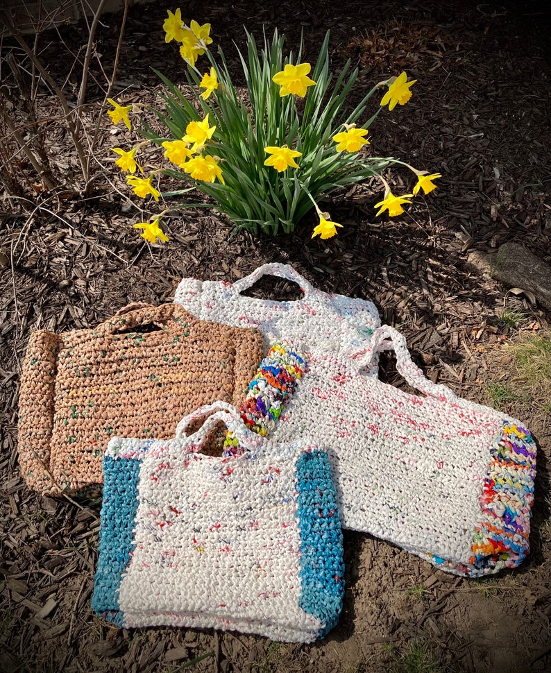 May include: Three crocheted bags made from recycled plastic. The bags are in different colors and sizes. The largest bag is white with colorful accents, the medium bag is brown, and the smallest bag is white with blue accents.