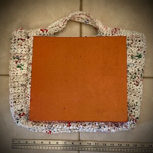 May include: A white crocheted bag with red, green, and blue accents. The bag is filled with a brown cardboard insert. A ruler is in the bottom of the image.