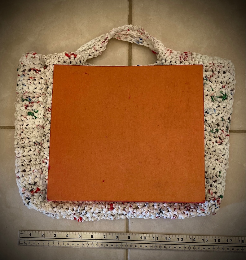 May include: A white crocheted bag with red, green, and blue accents. The bag is filled with a brown cardboard insert. A ruler is in the bottom of the image.