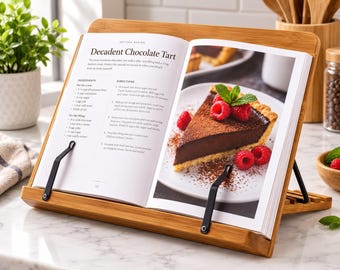 May include: A bamboo cookbook stand with two black metal page holders holding an open book titled Decadent Chocolate Tart. The page displays a recipe with ingredients and directions alongside a photograph of a chocolate tart with raspberries.