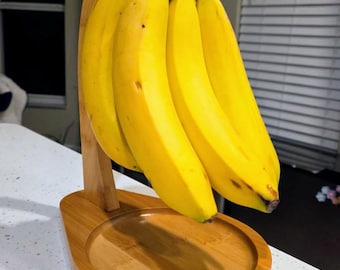May include: A light brown bamboo banana hanger featuring a curved vertical arm and a circular base. A bunch of four bright yellow bananas hangs from the top hook, suspended above the wooden tray, showcasing a natural wood grain finish.