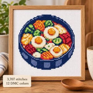 May include: A square cross-stitch embroidery in a light wood frame depicting a bowl of bibimbap with three fried eggs, green vegetables, red kimchi, and yellow ingredients. A small sign reads 3,707 stitches and 12 DMC colours in black text.