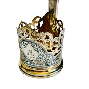 Könnte beinhalten: Ein metallener Teeglashalter mit einem filigranen oberen Rand und einem massiven Boden. Der Körper zeigt ein graviertes Blumenmuster, das eine zentrale Kartusche mit dem Buchstaben M und dem Datum 31.VIII.59 auf der Vorderseite umgibt.