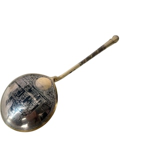 May include: A silver-toned metal spoon featuring a large, oval-shaped bowl with an intricate black engraved illustration of a building with domes and towers. The handle is slender, metallic, and decorated with a spiral pattern, ending in a rounded tip.