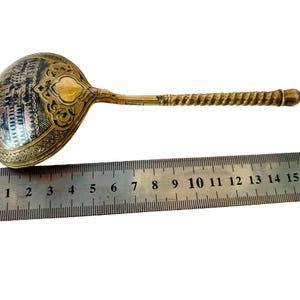 May include: A gold-toned metal ladle with a round bowl featuring an intricate engraved architectural scene and a twisted handle. A metal ruler is positioned underneath, showing the ladle measures approximately 18 cm in total length.