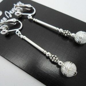 May include: A pair of silver clip-on earrings with a long, thin silver rod and a textured silver ball at the end. The earrings are on a black card with the text "Fashion Jewelry" written on it.