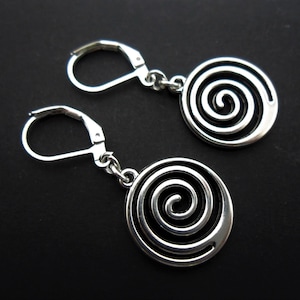 May include: Silver-toned earrings featuring a spiral design within a circular frame. The earrings have lever-back closures and hang from a small chain. The spiral design is a prominent feature, creating a modern and eye-catching look. The earrings are displayed against a dark background.