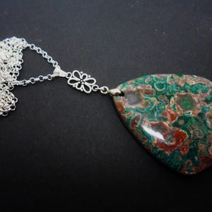 May include: A necklace featuring a large, teardrop-shaped polished stone pendant with a mottled pattern of deep green, brown, and reddish-orange colors. The pendant is attached to a silver-toned metal chain by a decorative floral-shaped link.