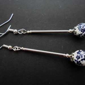 Puede incluir: Pendientes colgantes con cuentas estampadas con motivos florales blancos y azules. Los pendientes tienen ganchos de metal plateado y barras largas y delgadas. Las cuentas tienen forma ovalada y presentan un delicado diseño floral azul.