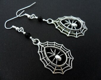 May include: Silver dangle earrings with a spider in a web design. The earrings have a black bead accent.