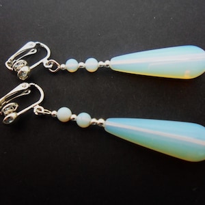 May include: A pair of silver-tone clip-on earrings featuring two small round opalescent beads and a long, smooth, teardrop-shaped opalescent pendant. The earrings have a metallic silver finish and a translucent, milky blue appearance with subtle light reflections.