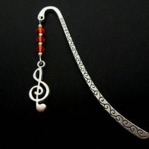 May include: Silver bookmark with a treble clef charm and orange beads. The bookmark has a decorative design of swirls along the edge.