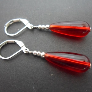 May include: A pair of drop earrings featuring teardrop-shaped, translucent red beads. The earrings have silver-coloured lever-back closures and small silver beads. The earrings are displayed on a dark surface.