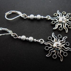 May include: Silver-toned dangle earrings featuring a sun design. Each earring has a lever-back closure, a chain of small beads, and a detailed sun pendant with a face in the center. The earrings are displayed against a dark background.