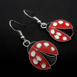 Può includere: Un paio di orecchini a coccinella. Ogni orecchino presenta un design a coccinella in smalto rosso con pois bianchi e dettagli neri. Gli orecchini hanno ganci per le orecchie color argento.