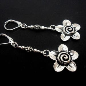 Può includere: Un paio di orecchini d'argento con un design a forma di fiore. Ogni orecchino presenta un fiore d'argento con un vortice al centro e una catena di perline d'argento.