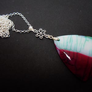 May include: A silver-tone chain necklace featuring a small, six-petaled flower connector attached to a large, triangular pendant. The pendant has a smooth, polished surface with a cracked pattern in white, teal, and deep magenta color sections.