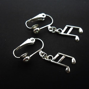 Op de afbeelding: Een paar zilveren clip-on oorbellen met een muzieknotenhanger. De oorbellen zijn gemaakt van metaal en hebben een eenvoudig ontwerp.